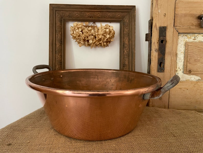 Puede incluir: Una olla grande de cobre con dos asas de hierro. La olla est&aacute; sobre una superficie de arpillera.