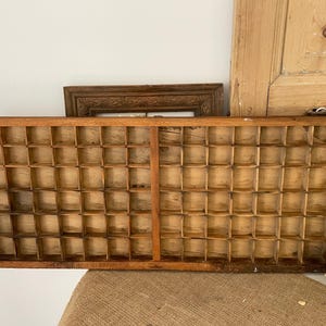 Könnte beinhalten: Ein rechteckiges Setzkasten-Tablett aus Holz mit einem Raster aus zahlreichen kleinen, offenen Fächern. Das Tablett ist aus hellbraunem Holz mit sichtbarer Maserung und einer verwitterten Oberfläche gefertigt und bietet eine strukturierte Anordnung.