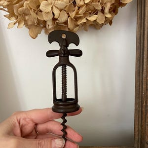 Könnte beinhalten: Ein dunkelbrauner Metallkorkenzieher mit einer Wendel und einem Griff. Der Korkenzieher wird vor einem Kranz aus getrockneten Blumen und einem Holzbilderrahmen gehalten. Der Korkenzieher ist ein Weinöffner im Vintage-Stil.