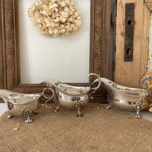 May include: Three silver gravy boats with ornate handles and feet are displayed on a burlap surface. The gravy boats have a reflective surface and are arranged in a row, showcasing their elegant design. The background includes a wooden frame and a dried floral arrangement.