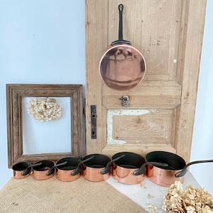 May include: A selection of copper pots and pans with black handles. A large copper pan hangs on a wooden door. A framed artwork and a burlap surface are also in the image. The pots and pans are of different sizes.