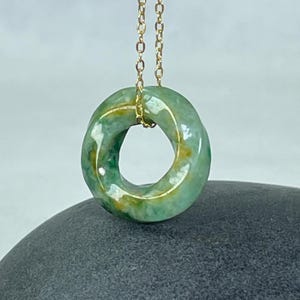 Peut inclure: Un collier pendentif composé d'une pierre circulaire en forme de beignet aux motifs verts et jaunes, suspendue à une fine chaîne en métal doré. La pierre présente une surface lisse et polie avec des variations de couleurs naturelles visibles.