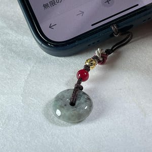 Puede incluir: Un dije de teléfono de jade con un cordón oscuro, cuentas rojas y detalles dorados, sujeto a un teléfono inteligente. El jade es de color verde claro y tiene forma circular. La pantalla del teléfono muestra texto japonés.