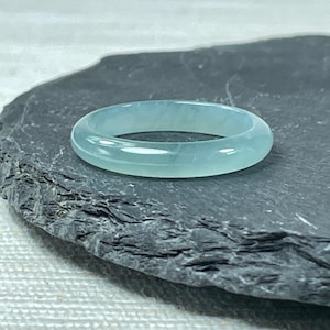 May include: A light blue jadeite bangle ring, with a smooth, polished finish. The ring is displayed on a dark gray slate surface.