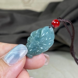 Könnte beinhalten: Ein Jade-Anhänger in Blattform mit einem detaillierten Design. Der Anhänger ist an einer braunen Schnur mit einer roten Perle befestigt.