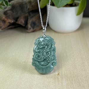 Op de afbeelding: Een jade hangerketting met een gedetailleerde drakengravure. De hanger is lichtgroen en hangt aan een zilveren ketting.