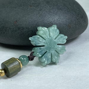 Puede incluir: Un colgante de jade con forma de flor con un diseño floral tallado, en tonos verdes y azules. El colgante está sujeto a un cordón marrón oscuro con cuentas de madera y doradas. El fondo presenta una piedra gris oscuro.