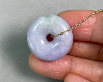 Puede incluir: Un colgante redondo de jade lavanda con una cadena de oro. El colgante tiene un agujero en el centro y una superficie pulida. El jade presenta una mezcla de tonos lavanda y verde.