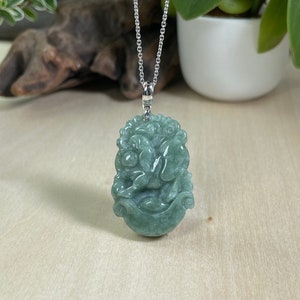 Puede incluir: Un collar con un colgante de jade verde tallado con una criatura mítica. El colgante está suspendido de una cadena de plata.