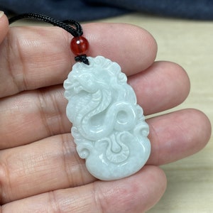May include: White jade pendant carved with a detailed dragon design. The pendant is attached to a black cord with a red bead.
