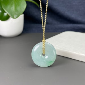 May include: A jade donut pendant necklace with a gold chain. The pendant is a pale green color with a circular shape and a hole in the center. The gold chain is delicate and hangs from the pendant.