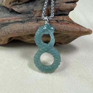 Op de afbeelding: Lichtblauwe jade hanger in de vorm van een oneindigheidssymbool, hangend aan een zilveren ketting. De hanger heeft een textuur en wordt gepresenteerd tegen een houten achtergrond.