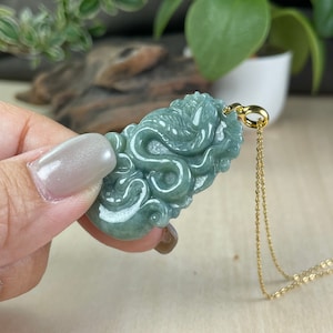 May include: A jade pendant carved in the shape of a dragon, with a gold chain. The pendant is a mix of green and white, with intricate details. The gold chain is delicate and thin, with a simple clasp.