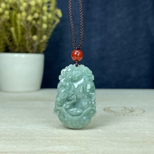 Puede incluir: Un collar con un colgante de jade verde tallado con una criatura mítica. El colgante está unido a un cordón marrón con una cuenta roja.
