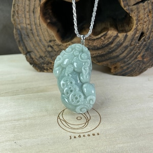 Könnte beinhalten: Ein Anhänger aus Jadegrün, der in Form eines Fabelwesens mit filigranen Details geschnitzt ist. Der Anhänger hängt an einer silbernen Kette.