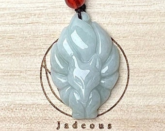 Op de afbeelding: Een lichtgroene jade hanger gegraveerd in de vorm van een negenstaartige vos, hangend aan een bruin koord met een rode kraal. De hanger ligt op een houten oppervlak met het woord "Jadecous" erop gegraveerd.