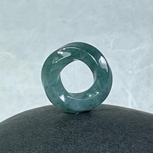May include: A jade donut pendant with a swirling design. The pendant is a translucent teal green color and has a polished finish. The circular shape is a classic design, often used in jewelry.