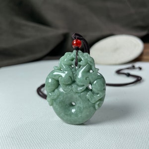 May include: A jade pendant necklace featuring a green jade carving of two mythical creatures, possibly dragons or phoenixes, intertwined around a circular jade disc. The pendant is strung on a brown cord with a red bead.