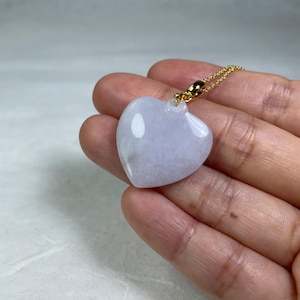 May include: A heart-shaped pendant necklace. The pendant is a pale lavender color with a polished surface. It hangs from a delicate gold chain. The necklace is a piece of jewelry.