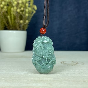 May include: A jade green pendant carved in the shape of a rooster, with intricate details and a brown cord necklace. The pendant is a traditional Chinese symbol of good luck and prosperity.