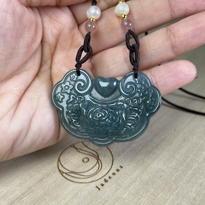 May include: A jade pendant necklace with a carved floral design. The pendant is green and has a heart shape in the center. The necklace is made of black cord and has white and pink beads.