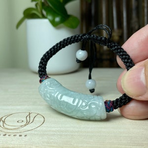 Op de afbeelding: Een groene jade armband met ingewikkelde gravures, bestaande uit een zwart gevlochten koord met witte kralen en een knoopsluiting.