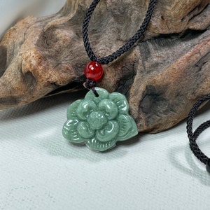 May include: A green jade flower pendant on a black cord necklace. The pendant is carved in the shape of a flower with a detailed centre.