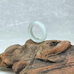 May include: A light green jade ring. The ring is a solid band with a smooth, polished surface. The jade has a translucent quality, with subtle variations in color. The ring is displayed on a piece of weathered wood.