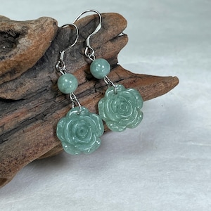 May include: A pair of jade earrings featuring a floral rose design. Each earring has a silver hook, a round jade bead, and a carved jade rose pendant. The earrings are a light green color and are displayed on a piece of driftwood.