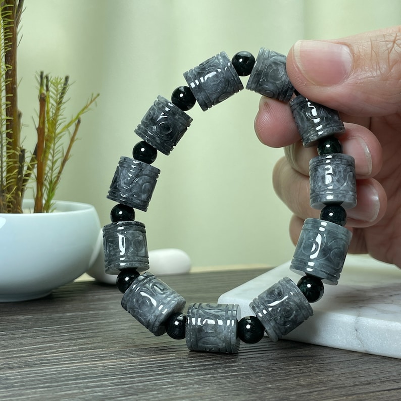 Puede incluir: Un brazalete de cuentas grises y negras. El brazalete presenta cuentas cil&iacute;ndricas grises con dise&ntilde;os tallados, intercaladas con peque&ntilde;as cuentas negras redondas. El brazalete se muestra sobre un fondo neutro.