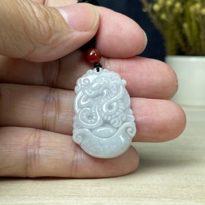 May include: White jade pendant carved with a detailed dragon design. The pendant is oval shaped and has a smooth, polished finish.