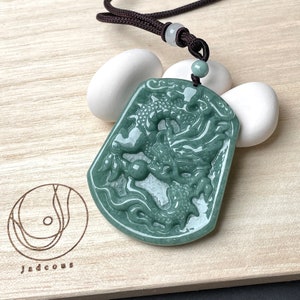 Könnte beinhalten: Ein Anhänger aus grünem Jade mit einer detaillierten Schnitzerei eines Drachen. Der Anhänger ist an einer braunen Schnur mit einer kleinen Jadeperle befestigt.