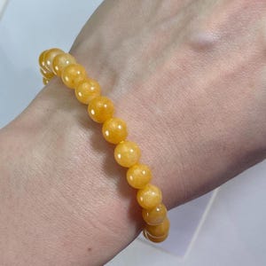 Puede incluir: Una pulsera con cuentas esféricas de color amarillo dorado. Las cuentas están ensartadas, formando un bucle continuo. La pulsera se muestra en una muñeca, con las cuentas en contacto con la piel. Las cuentas tienen un aspecto liso y pulido.