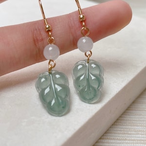 Könnte beinhalten: Ein Paar Jade-Blattohrringe mit einem geschnitzten Design. Die Ohrringe sind aus grünem Jade und haben einen goldenen Haken. Jeder Ohrring hat eine kleine weiße Perle über dem Jadeblatt.