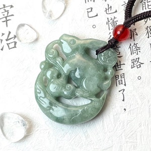 May include: A jade green pendant featuring a carved rabbit design. The pendant is attached to a black cord with a red bead.