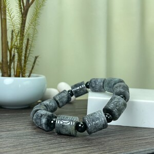 Puede incluir: Una pulsera de cuentas grises con cuentas cil&iacute;ndricas y cuentas negras. La pulsera est&aacute; hecha de un material de piedra y se exhibe sobre una superficie de madera.