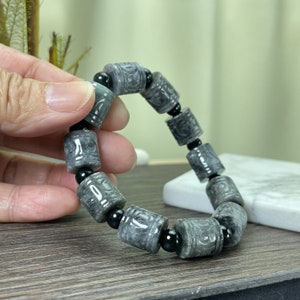 Puede incluir: Pulsera de jade gris con cuentas cil&iacute;ndricas con patrones tallados, intercaladas con cuentas negras. La pulsera se muestra, mostrando los intrincados detalles y las variaciones naturales de la piedra.