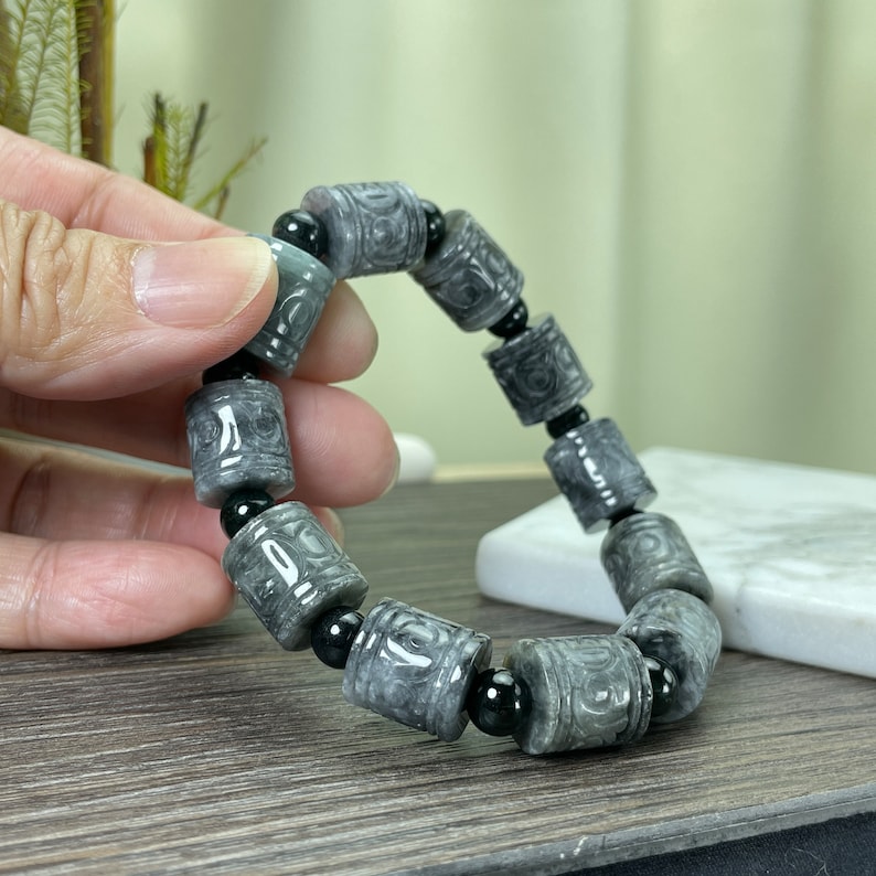 Puede incluir: Pulsera de jade gris con cuentas cil&iacute;ndricas con patrones tallados, intercaladas con cuentas negras. La pulsera se muestra, mostrando los intrincados detalles y las variaciones naturales de la piedra.