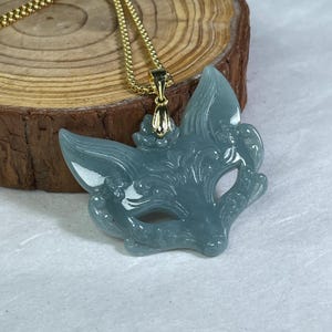Puede incluir: Un collar con colgante que presenta una máscara de zorro azul claro tallada. La máscara está suspendida de una cadena dorada. El colgante tiene detalles intrincados y se muestra sobre una superficie de madera.