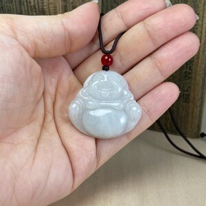 May include: A white jade pendant carved in the shape of a smiling Buddha, hanging from a black cord with a red bead. The pendant is about 2 inches tall and 1.5 inches wide.