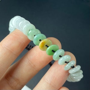 以下が含まれることがあります： 緑、黄色、白の翡翠ビーズがグラデーションになった翡翠のブレスレット。ビーズは黒い紐に通されています。