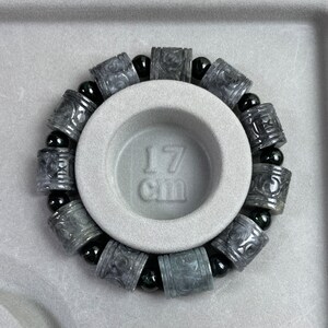 Puede incluir: Una pulsera gris con cuentas cil&iacute;ndricas talladas y cuentas esf&eacute;ricas negras. La pulsera se muestra sobre una superficie gris con una hendidura circular. El texto "17 cm" es visible.