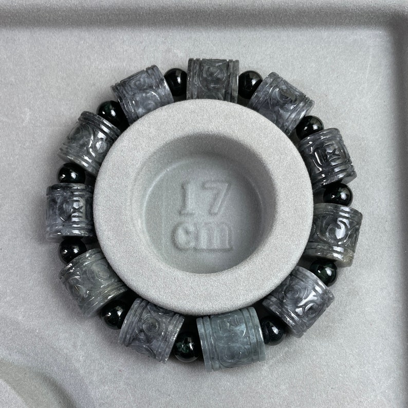 Puede incluir: Una pulsera gris con cuentas cil&iacute;ndricas talladas y cuentas esf&eacute;ricas negras. La pulsera se muestra sobre una superficie gris con una hendidura circular. El texto "17 cm" es visible.