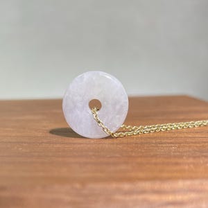 Könnte beinhalten: Halskette mit einem hellrosa Jade-Donut-Anhänger und einer goldenen Kette. Der runde Anhänger hat ein Loch in der Mitte. Die Halskette wird auf einer Holzoberfläche präsentiert.