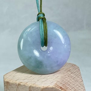 May include: A round, polished jade pendant with a hole in the center. The pendant is a mix of pale green and lavender hues, suspended by a green and teal cord. The pendant rests on a small wooden block.