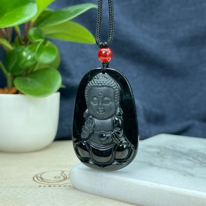 May include: Black obsidian pendant necklace featuring a carved Buddha figurine. The pendant is oval-shaped and has a black cord.