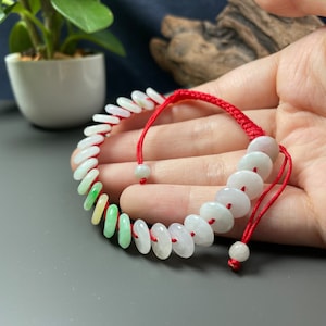 Könnte beinhalten: Ein Jade-Armband mit einem roten Schnallenverschluss. Das Armband besteht aus runden Jadeperlen mit einem Farbverlauf von Grün zu Weiß. Die Perlen sind auf einer roten Schnur aufgefädelt, die am Ende mit einem Knoten befestigt ist.