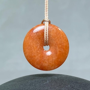 May include: A circular, orange-toned stone pendant with a smooth, polished surface and a central hole, suspended by a thin, light-colored braided cord. The stone features subtle internal variations in color and texture throughout its round form.