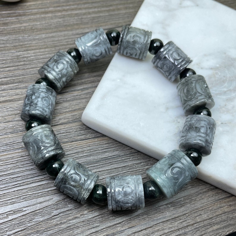 Puede incluir: Una pulsera gris con cuentas cil&iacute;ndricas talladas con un dise&ntilde;o en espiral. La pulsera est&aacute; ensartada con cuentas negras entre cada cuenta tallada. La pulsera se exhibe sobre una superficie de m&aacute;rmol blanco.