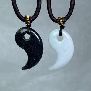 Pode incluir: Dois pingentes Yin Yang combinando, um preto e outro branco. Cada pingente é suspenso por um cordão preto com detalhes dourados. Os pingentes são lisos e polidos, com formato curvo e um furo na parte superior.